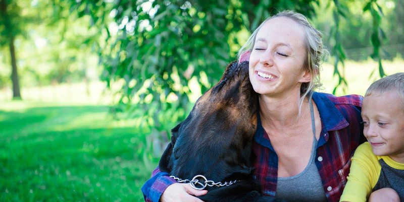A woman hugging a dog and child in a green backyard
